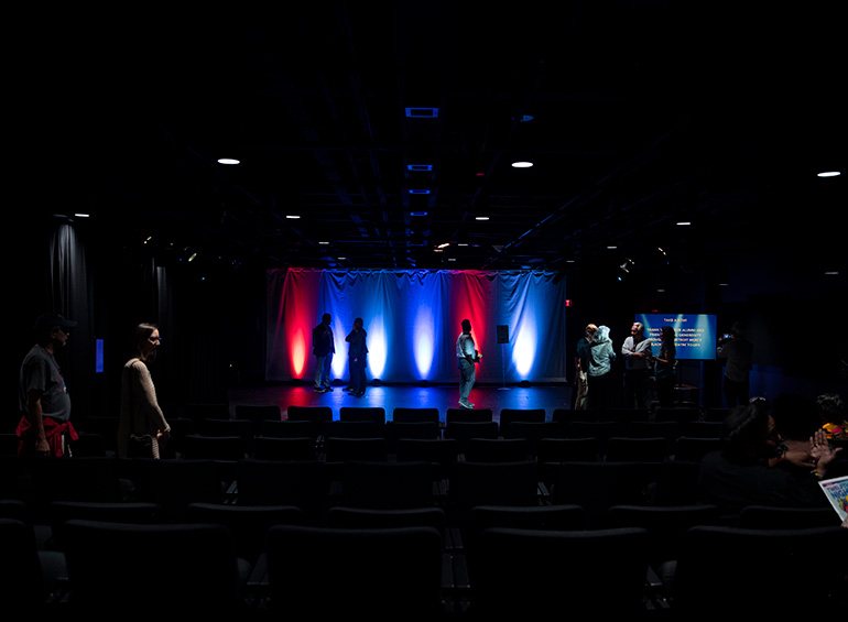 The dark Detroit Mercy Black Box Theatre, with people chatting on stage in front of a curtain lit red and blue