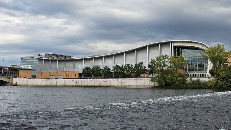 DeVos Place, with its wave-shaped roof and glass end wall, on the bank of the Grand River under a cloudy sky