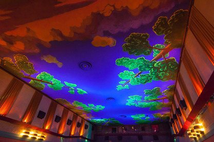 The auditorium ceiling seen from below, a painted sky of clouds and leafy trees glowing in blue and purple light