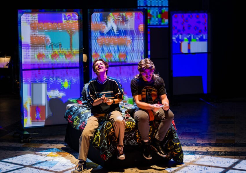 Two young actors laugh while sitting on a bed, with colorful video-game screens glowing behind them on stage