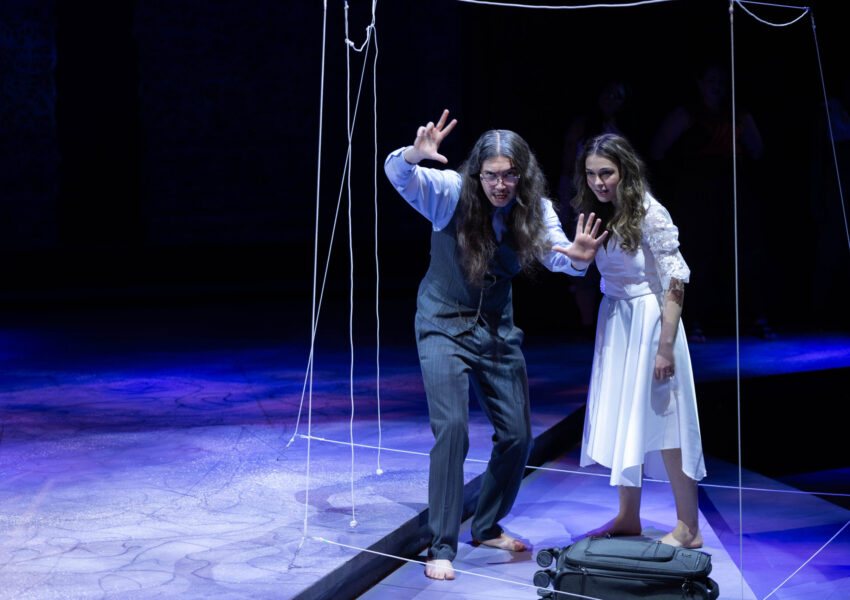 Two barefoot actors lean forward with outstretched hands on a violet-lit stage framed by taut ropes