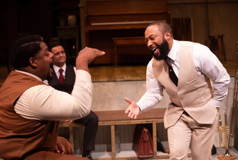 Two actors in vests laugh and gesture on a set with an old upright piano and wooden floor while a third sits on a bench behind them