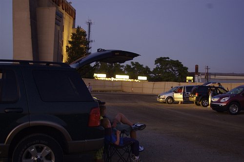 Cars and a viewer in a camp chair at dusk in the drive-in lot, with the tall screen tower on the left
