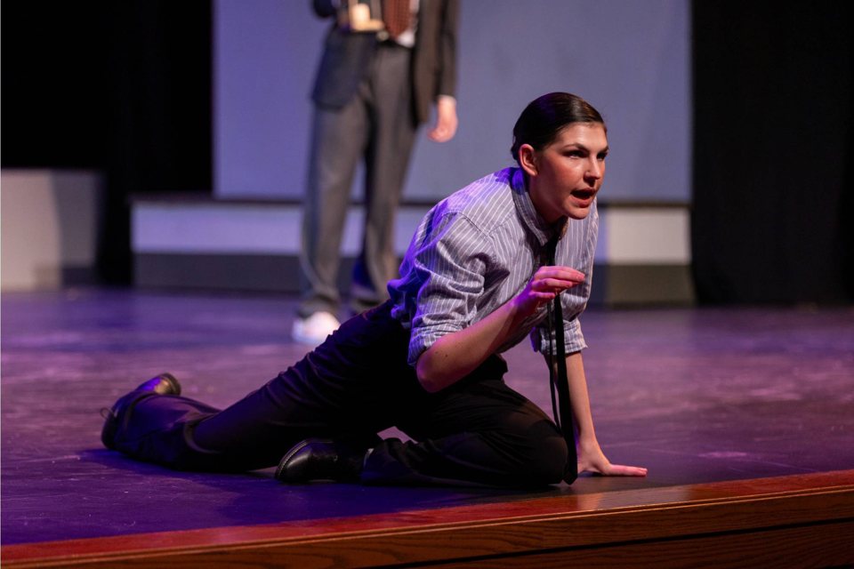 An actress in a striped shirt and loose tie kneels at the edge of the stage, with a suited performer standing behind her