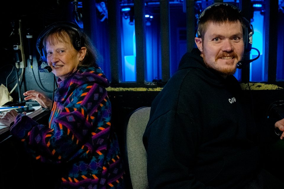 Two crew members in headsets smile in the control booth, with the stage glowing blue behind them