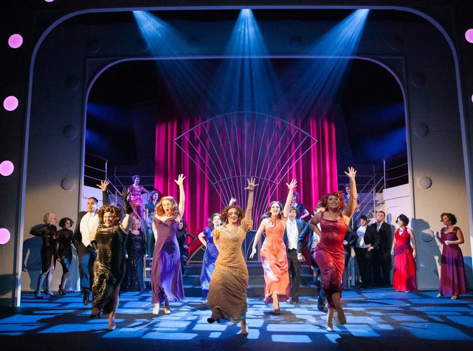The Anything Goes cast in evening gowns dancing with raised arms on a stage lit in blue
