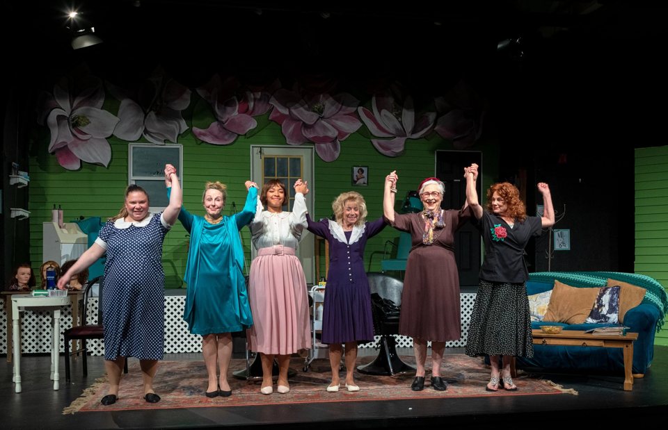 Six costumed actresses join raised hands for a curtain call in front of a green set painted with magnolias
