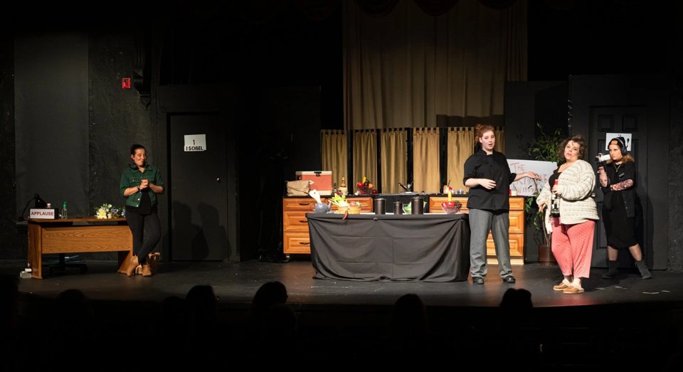 A TV cooking-show kitchen set on a dark stage, with four actors mid-scene