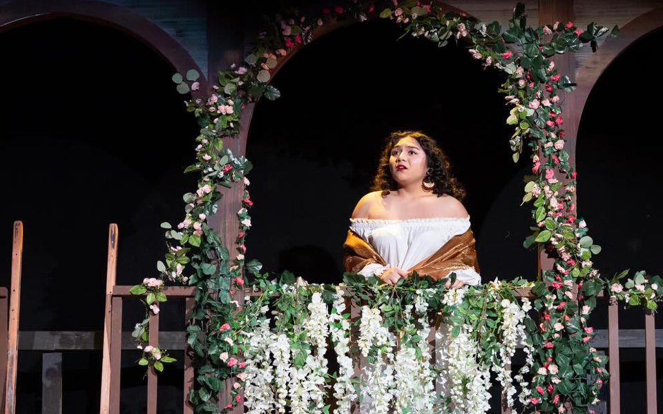An actress in a white blouse on a balcony draped with flower garlands, in front of a dark arched set