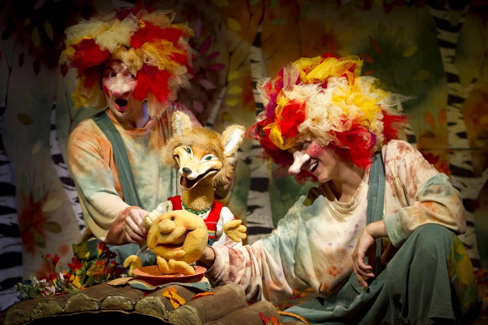Two puppeteers in colourful tulle wigs with a fox puppet and a Kolobok puppet on an autumn forest set