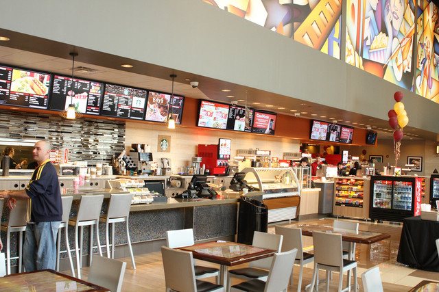 A modern cinema concession area with menu screens, high bar stools and a colorful mural below the ceiling
