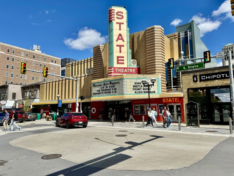 A State Theatre függőleges STATE felirata és márkiza napsütésben a State Street sarkán, előtte járókelőkkel