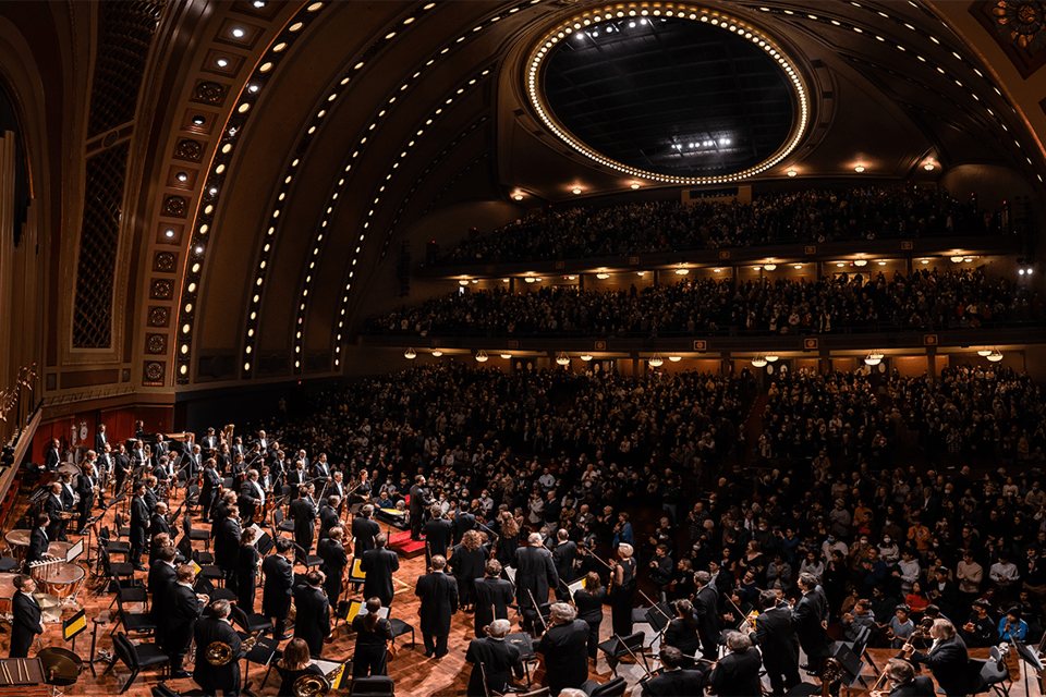 Teltházas Hill Auditorium: a zenekar állva fogadja a tapsot, fölötte az ívelt, fénysorokkal szegélyezett mennyezet és az erkélyek
