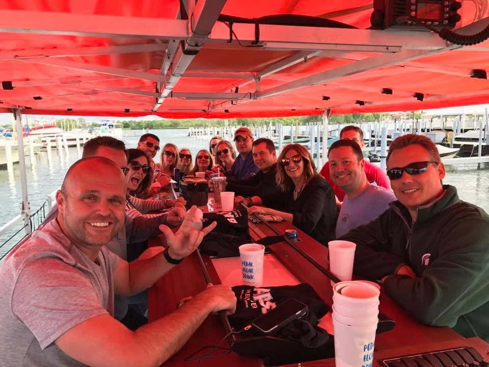 A group of friends sits around the pedal bar boat's counter under its red canopy
