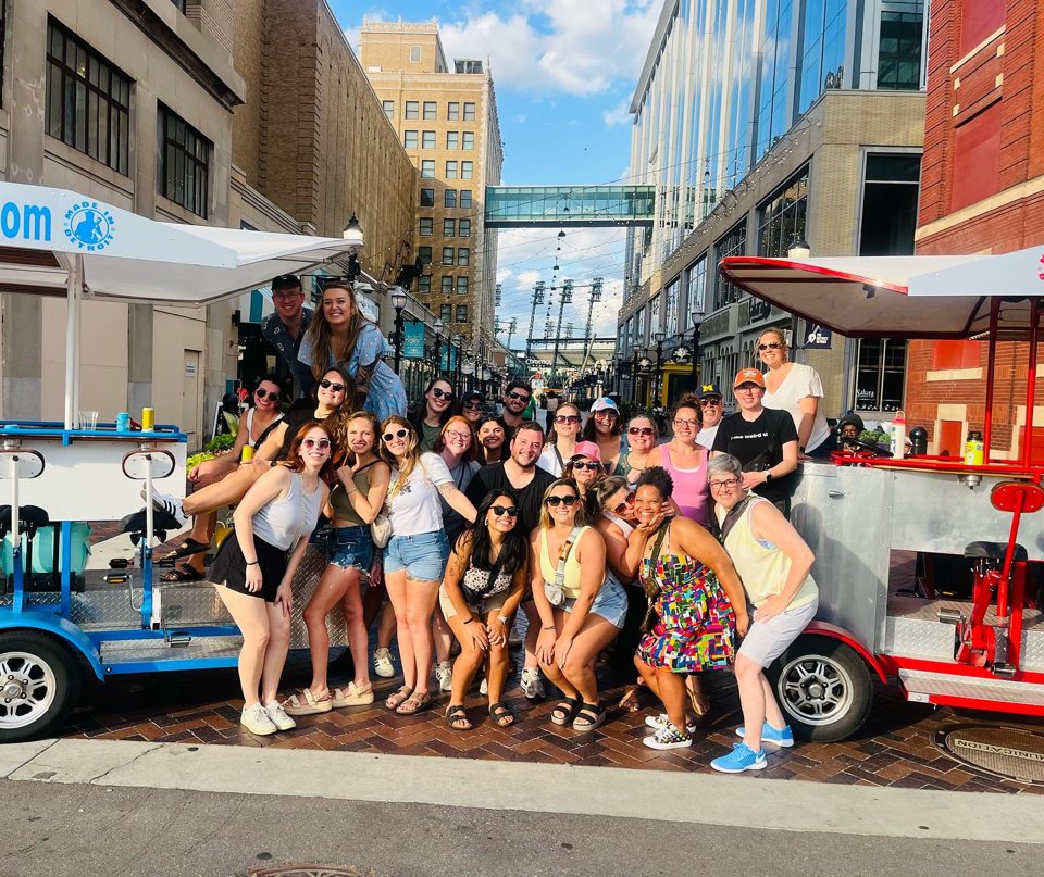 A large group stands between two pedal pubs in downtown Detroit, with Comerica Park behind them