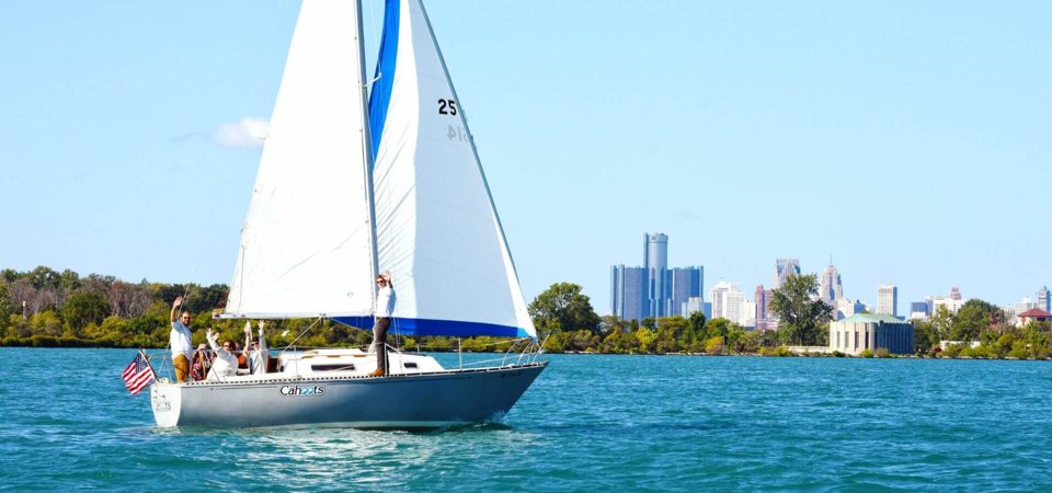 A sailboat glides across the water as passengers wave, with Detroit's Renaissance Center skyline behind
