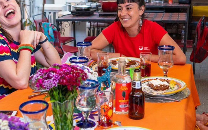 Vendégek nevetve ülnek egy kültéri asztalnál étellel és itallal, piros pólós túravezetővel
