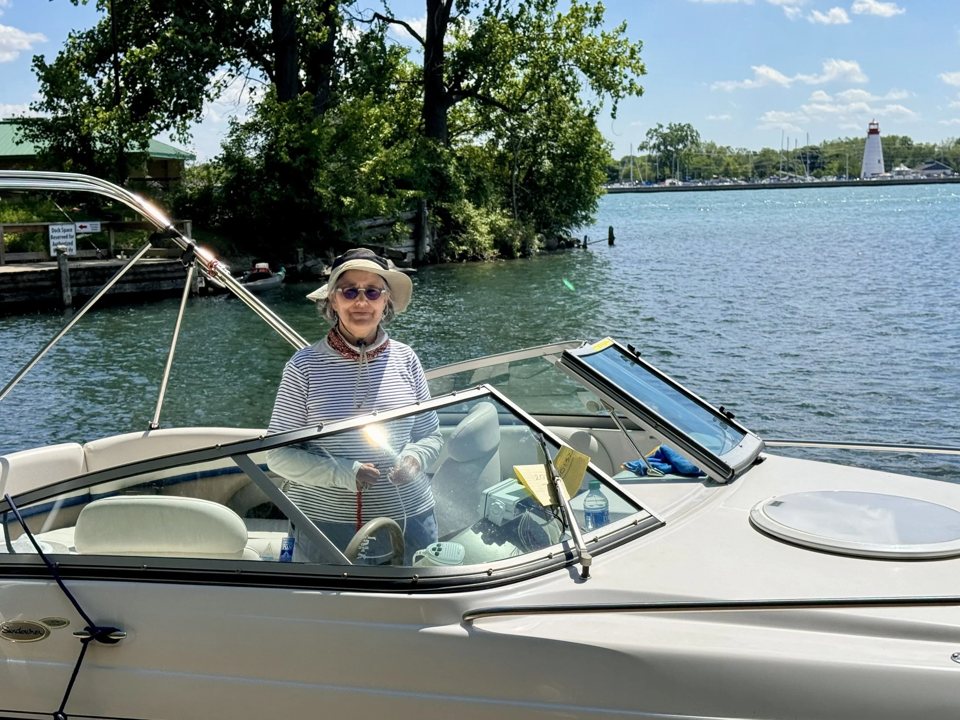 A woman in a sun hat drives the white motorboat on the river, with a lighthouse in the background