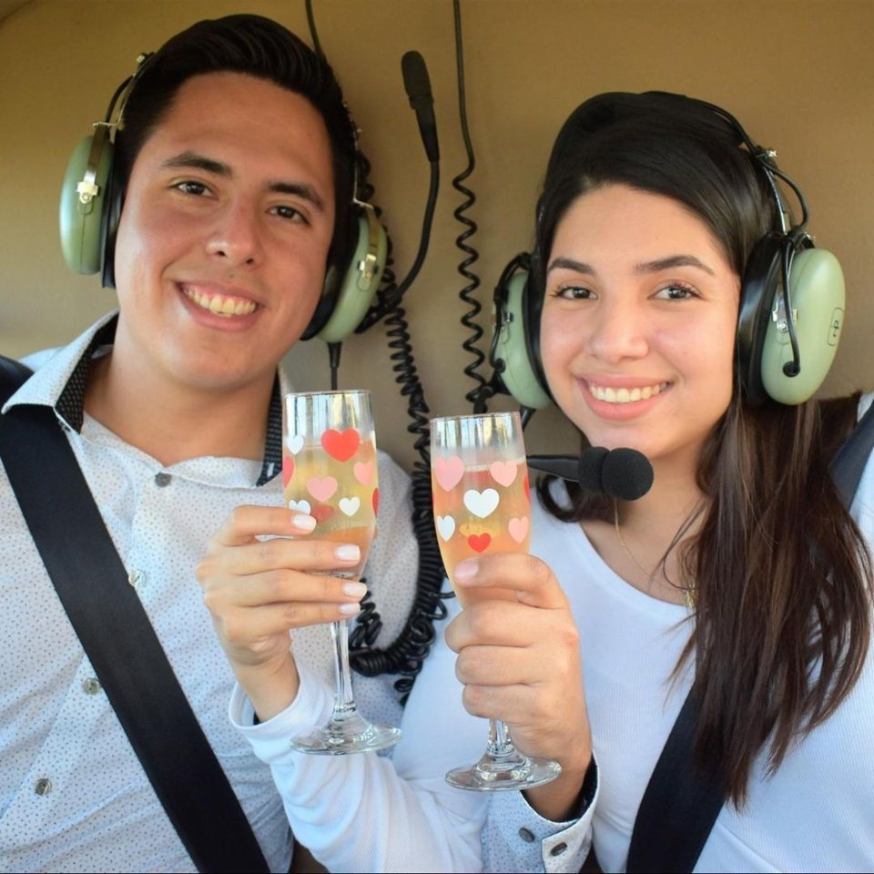 A young couple toasts with glasses of sparkling wine inside the helicopter, wearing headsets