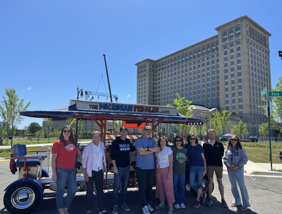 Csoport áll a Michigan Pedaler sörbiciklije előtt, mögöttük a Michigan Central pályaudvar